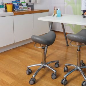 Praxishocker Pascals Junior 3D in Platin mit weißer Ziernaht. Mit silberner Ringauslösung und Basis Fußkreuz sowie grauen Medi Rollen und 3D Kippmechanik